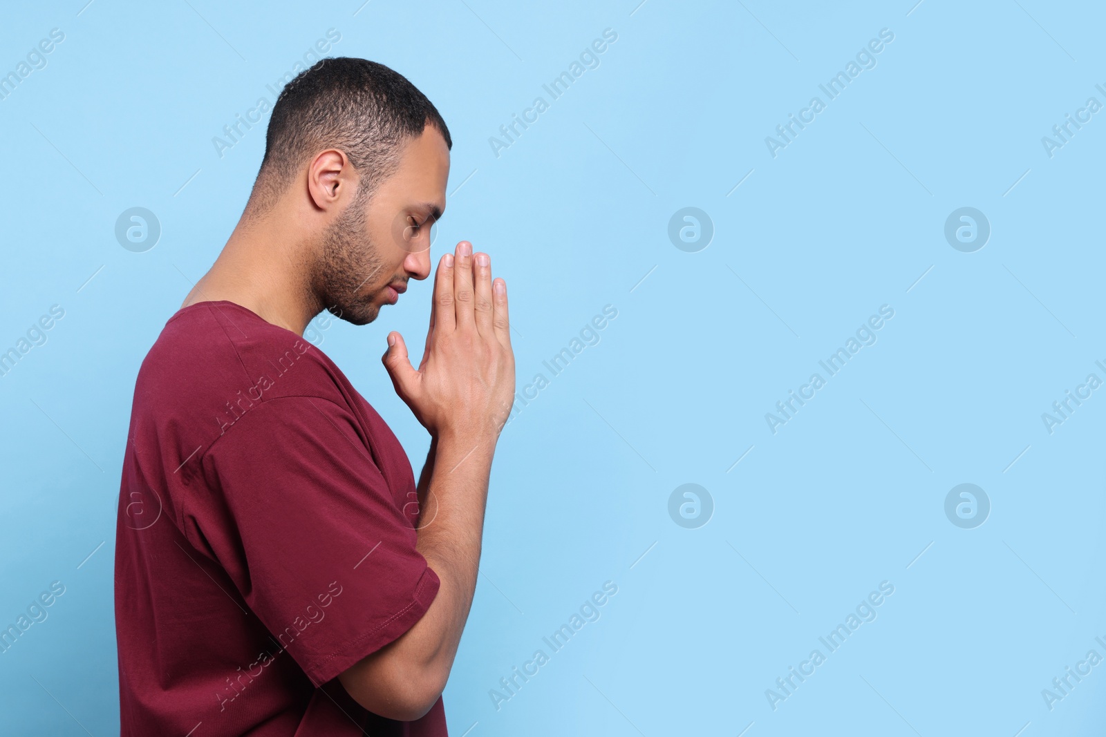 Photo of African American man with clasped hands praying to God on light blue background. Space for text