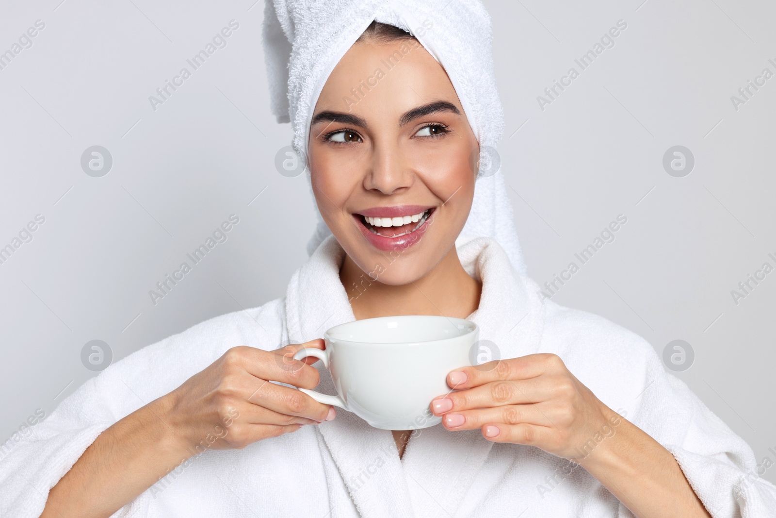 Photo of Young woman in bathrobe with towel and cup of drink on light grey background. Spa treatment