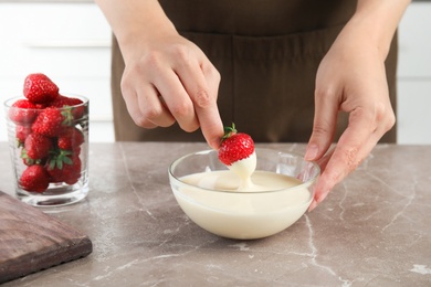 Photo of Woman dipping ripe strawberry into bowl with white melted chocolate on table