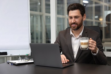 Photo of Man with cup of coffee working on laptop at black desk in office. Space for text