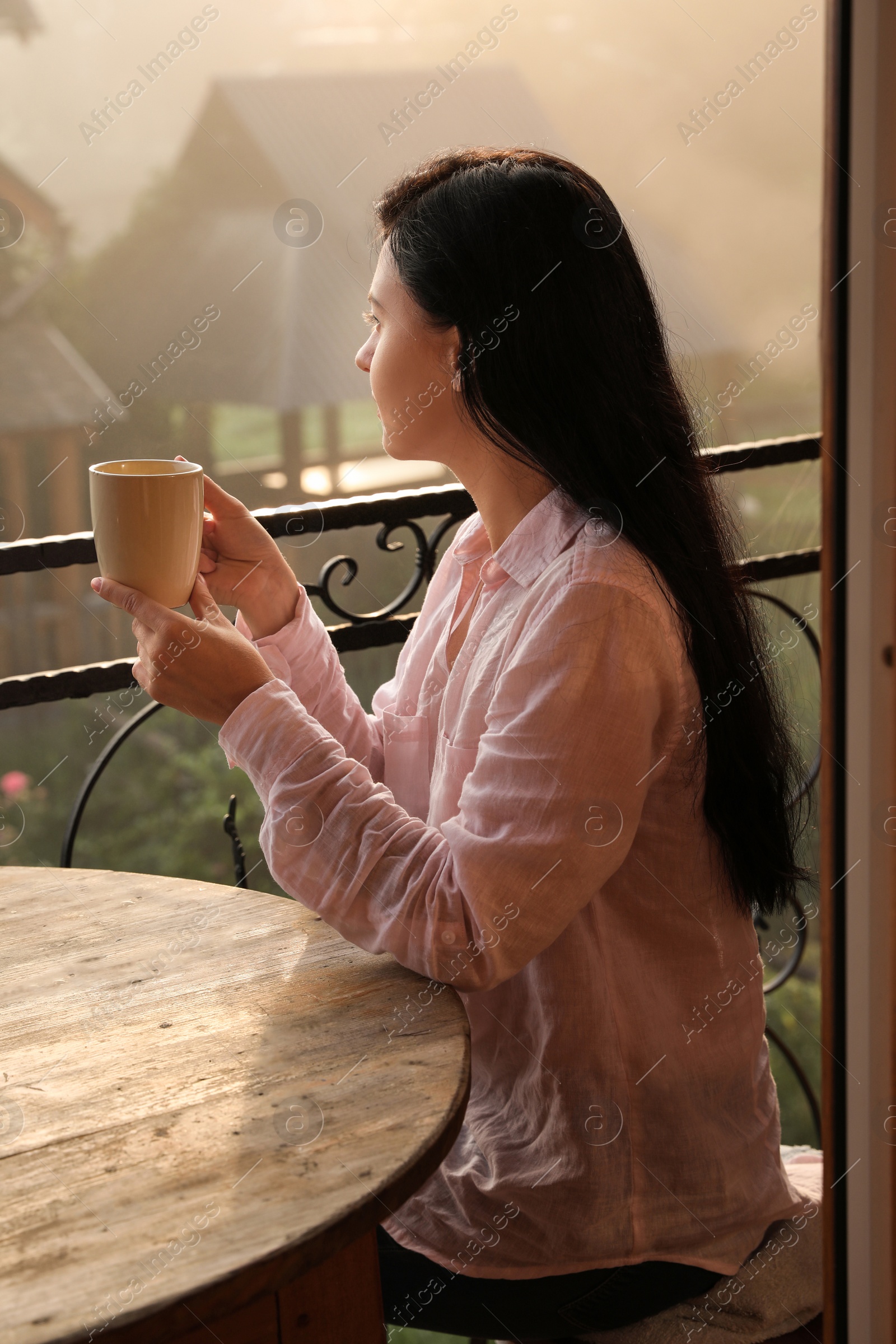 Photo of Young woman sitting at table on balcony with cup of tea in morning