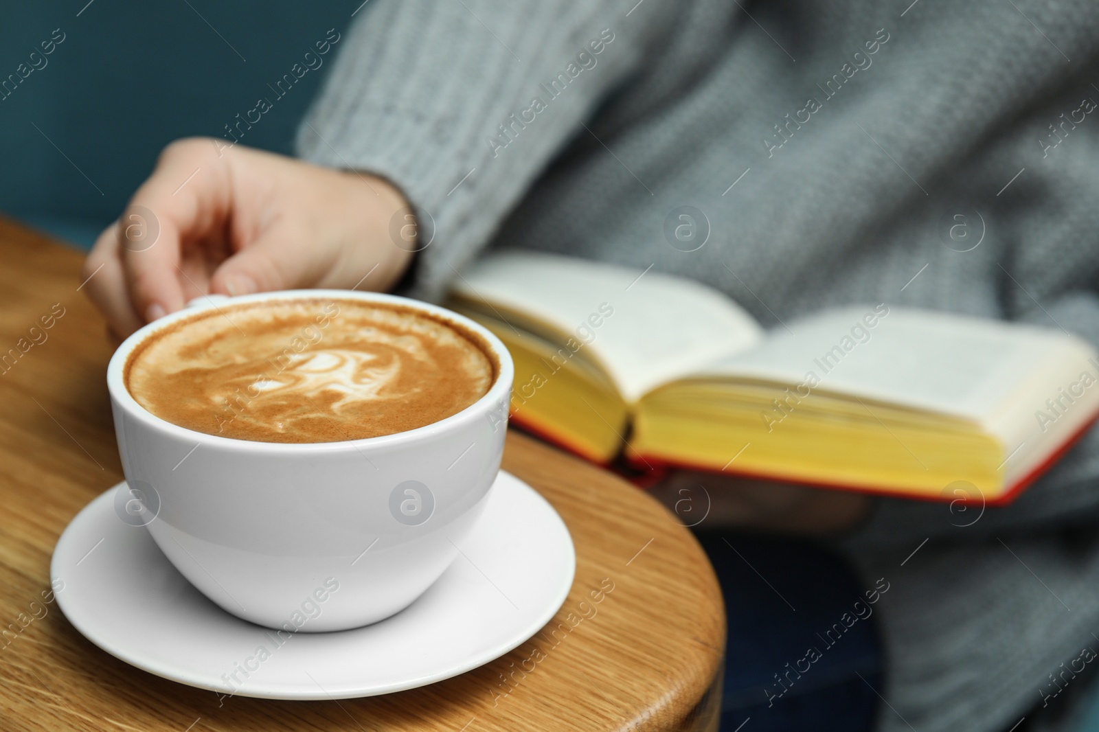 Photo of Woman reading book at table, focus on cup of coffee