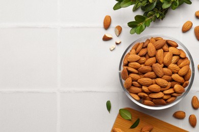 Bowl of delicious almonds and fresh leaves on white tiled table, flat lay. Space for text