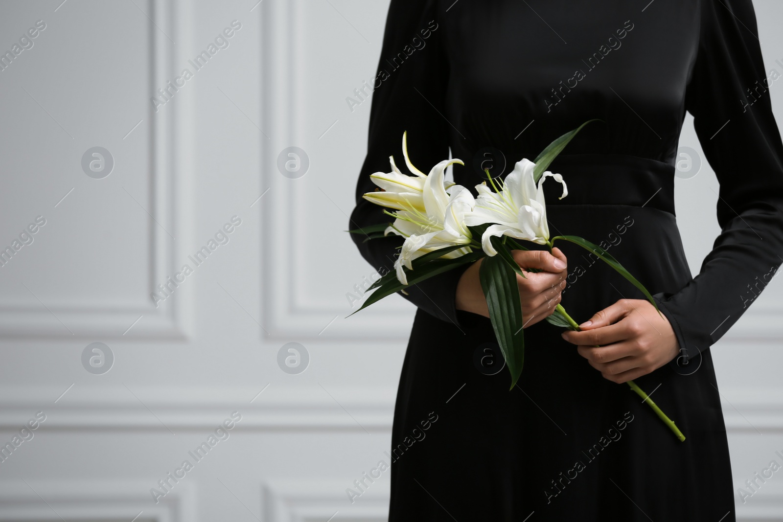 Photo of Woman with lilies near white wall, closeup and space for text. Funeral ceremony