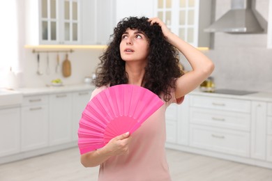Photo of Young woman waving pink hand fan to cool herself in kitchen
