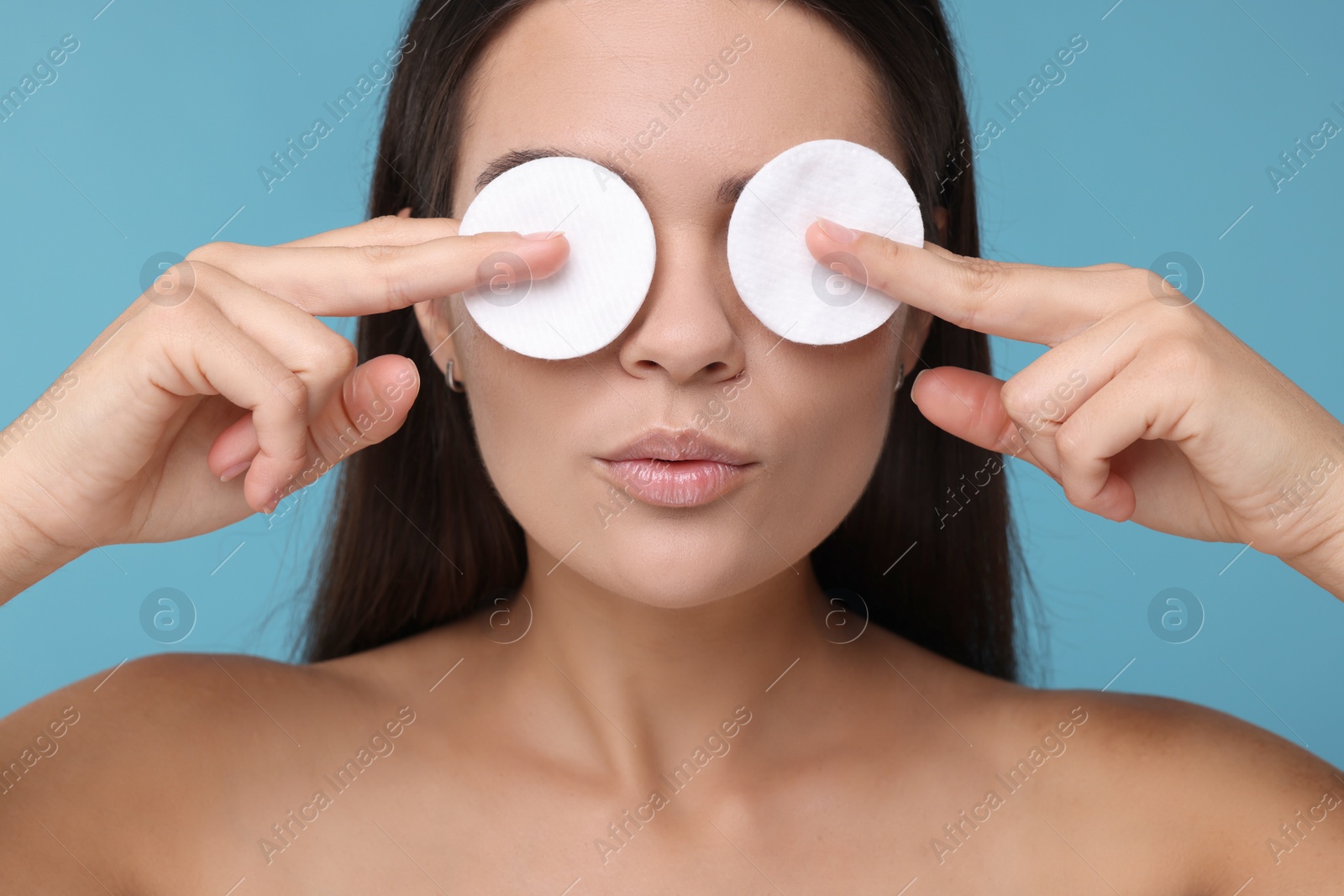 Photo of Young woman covering her eyes with cotton pads on light blue background, closeup