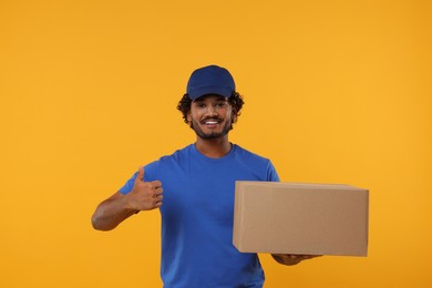 Photo of Happy young courier with parcel showing thumb up on orange background