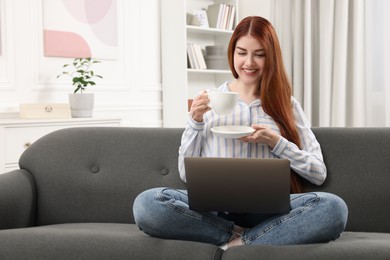 Photo of Happy woman with cup of drink using laptop on couch in room