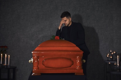 Sad young man near funeral casket with red rose in chapel