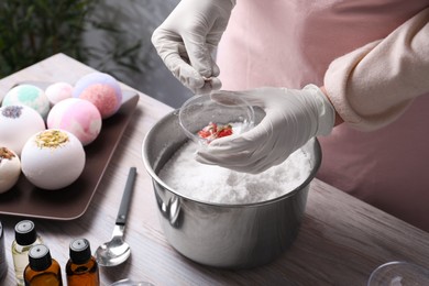 Photo of Woman in gloves filling bath bomb mold with dried flowers at wooden table indoors, closeup