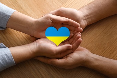Image of Stop war in Ukraine. Man and woman holding heart shaped symbol with colors of Ukrainian flag in hands at wooden table, closeup