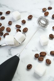 Photo of Black milk frother wand, sugar cubes and coffee beans on white marble table, closeup