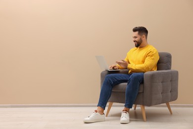 Handsome man with laptop sitting in armchair near beige wall indoors, space for text