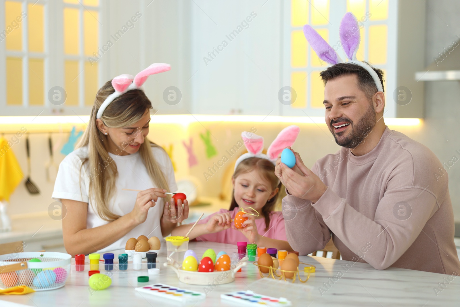 Photo of Easter celebration. Happy family with bunny ears painting eggs at white marble table in kitchen