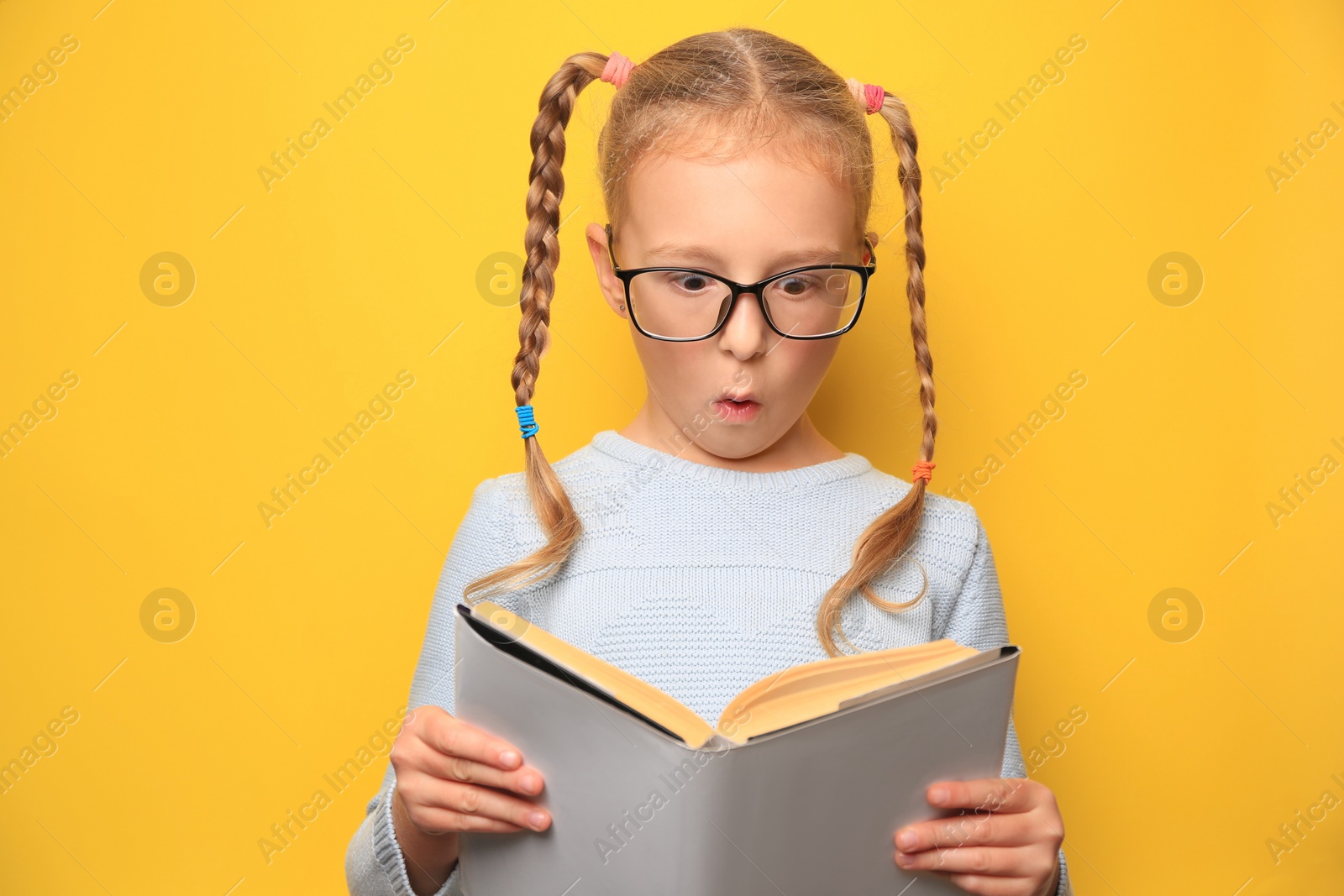 Photo of Cute little girl in glasses reading textbook on yellow background