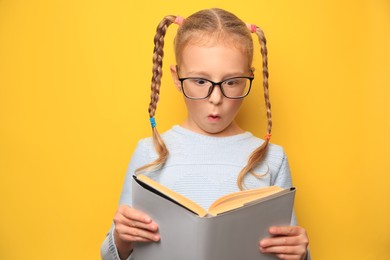 Photo of Cute little girl in glasses reading textbook on yellow background