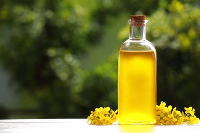 Photo of Rapeseed oil in glass bottle and yellow flowers on white wooden table outdoors, space for text
