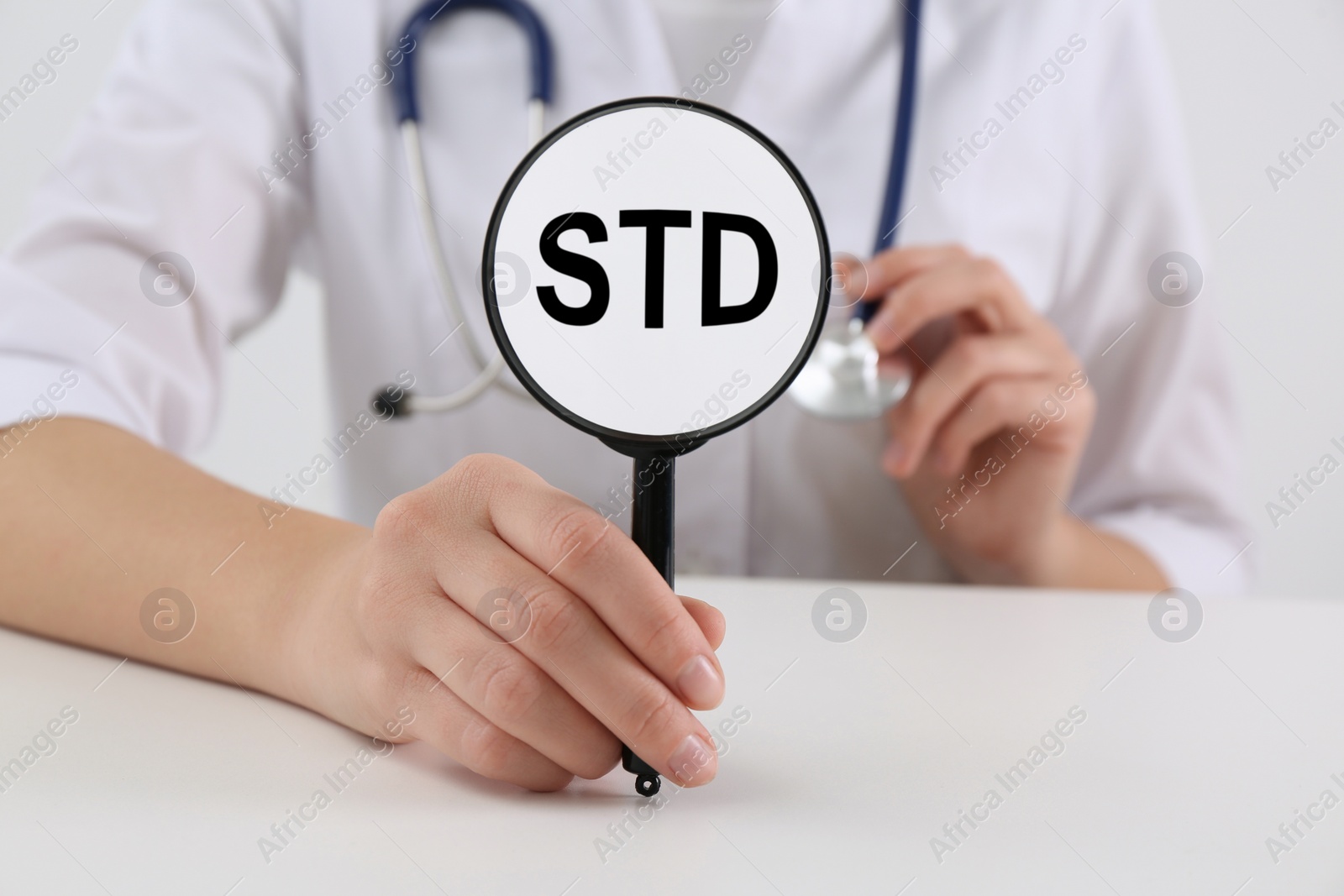 Photo of Doctor holding magnifying glass with abbreviation STD at white table, closeup