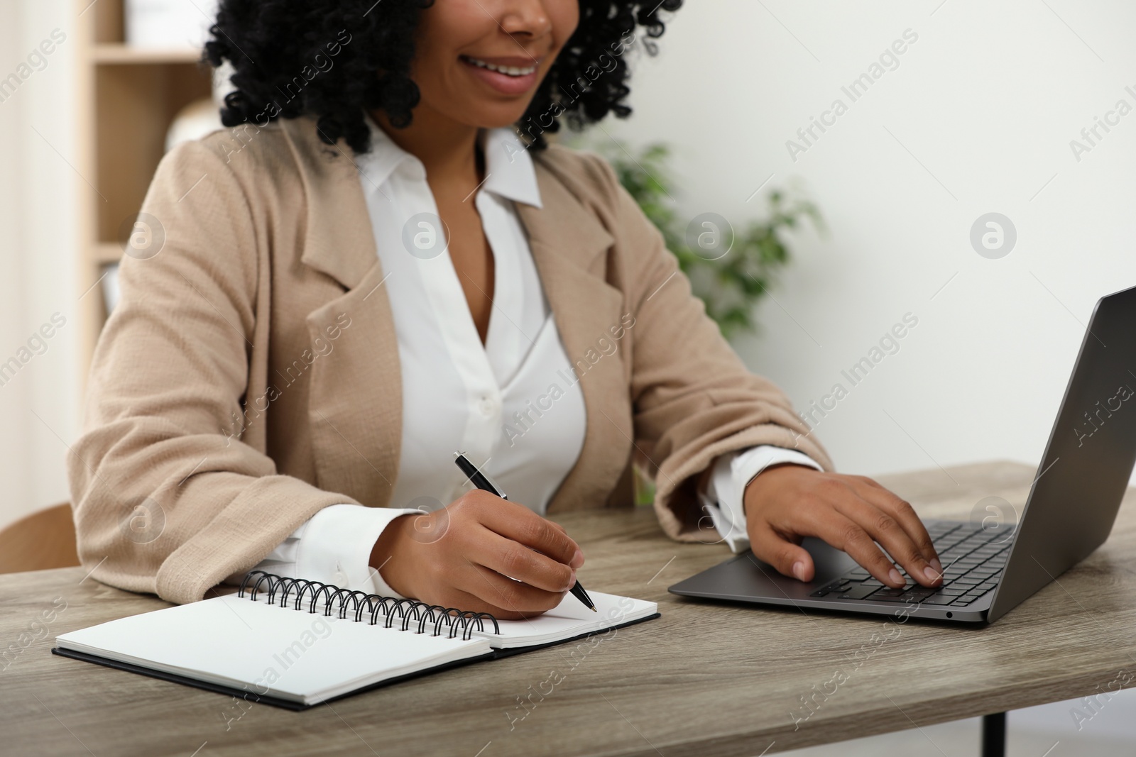 Photo of Woman writing notes while using laptop at wooden desk indoors, closeup