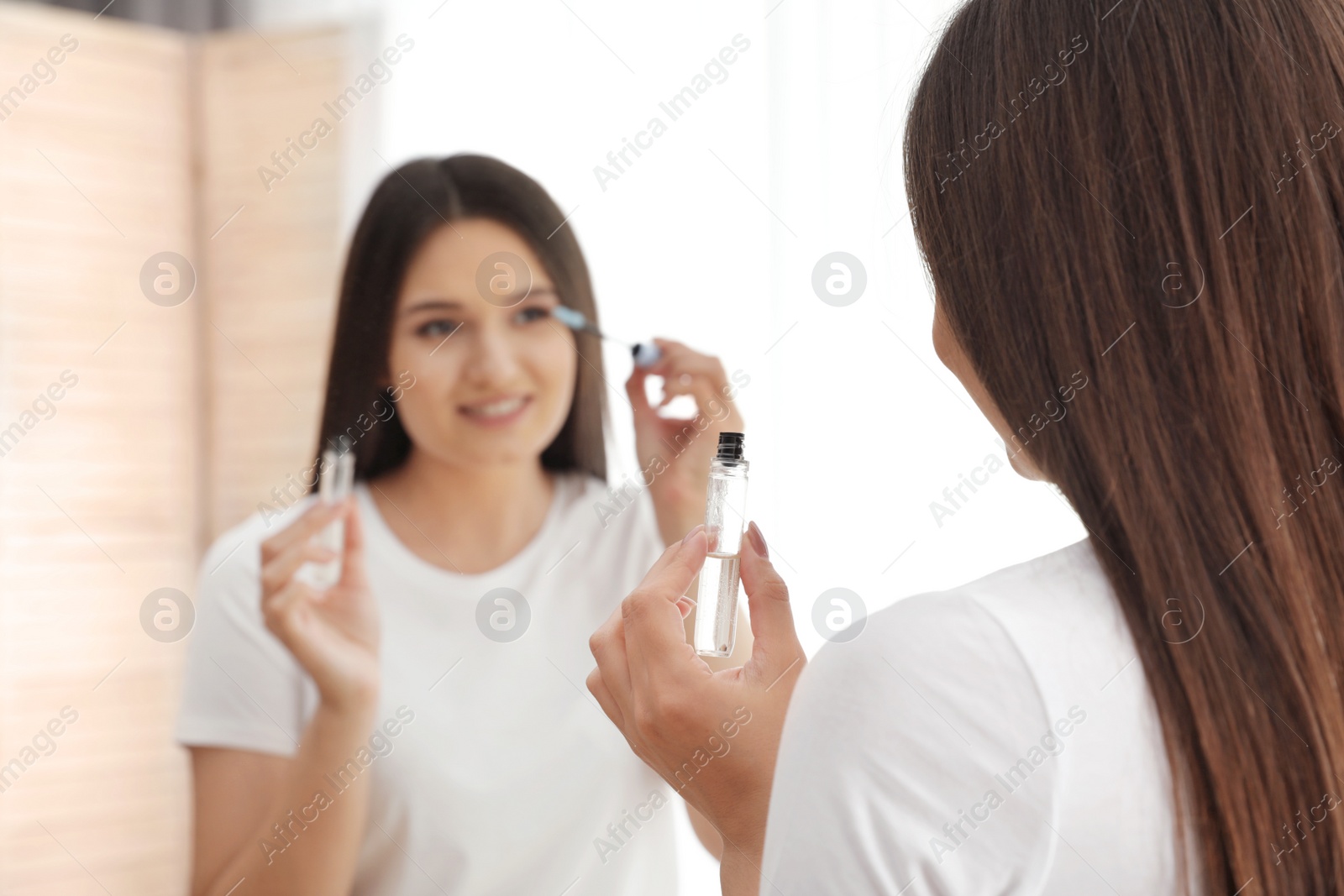 Photo of Beautiful woman applying oil onto her eyelashes near mirror indoors