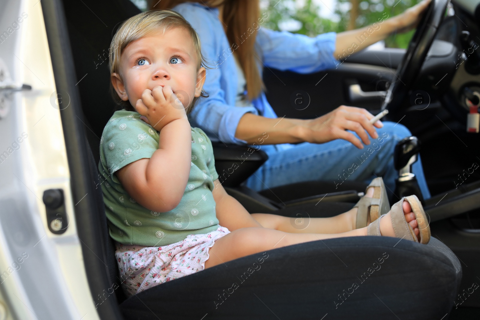 Photo of Mother with cigarette and child in car, focus on little girl. Don't smoke near kids