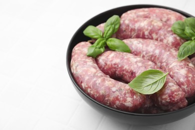 Photo of Raw homemade sausages and basil leaves in bowl on white tiled table, closeup. Space for text
