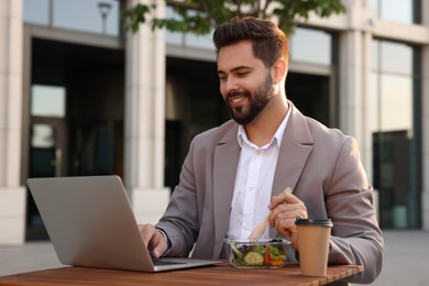 Photo of Happy businessman using laptop during lunch at wooden table outdoors