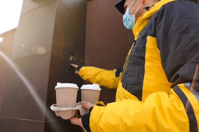 Photo of Courier in medical mask holding takeaway drinks and ringing doorbell outdoors, focus on paper cups. Delivery service during quarantine due to Covid-19 outbreak