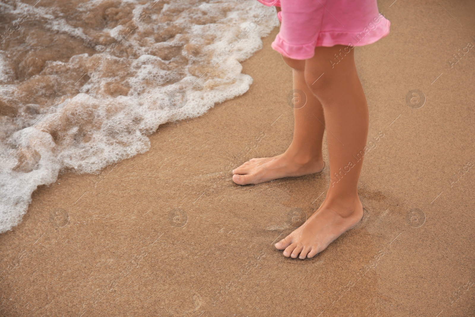 Photo of Little girl standing on sandy beach near sea, closeup. Space for text