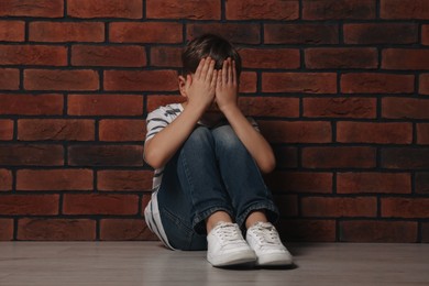 Photo of Upset boy sitting on floor near brick wall. Children's bullying