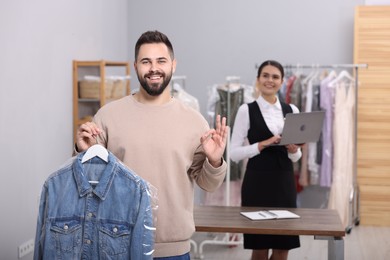 Photo of Dry-cleaning service. Happy man holding hanger with denim jacket and showing ok gesture indoors. Worker using laptop at workplace