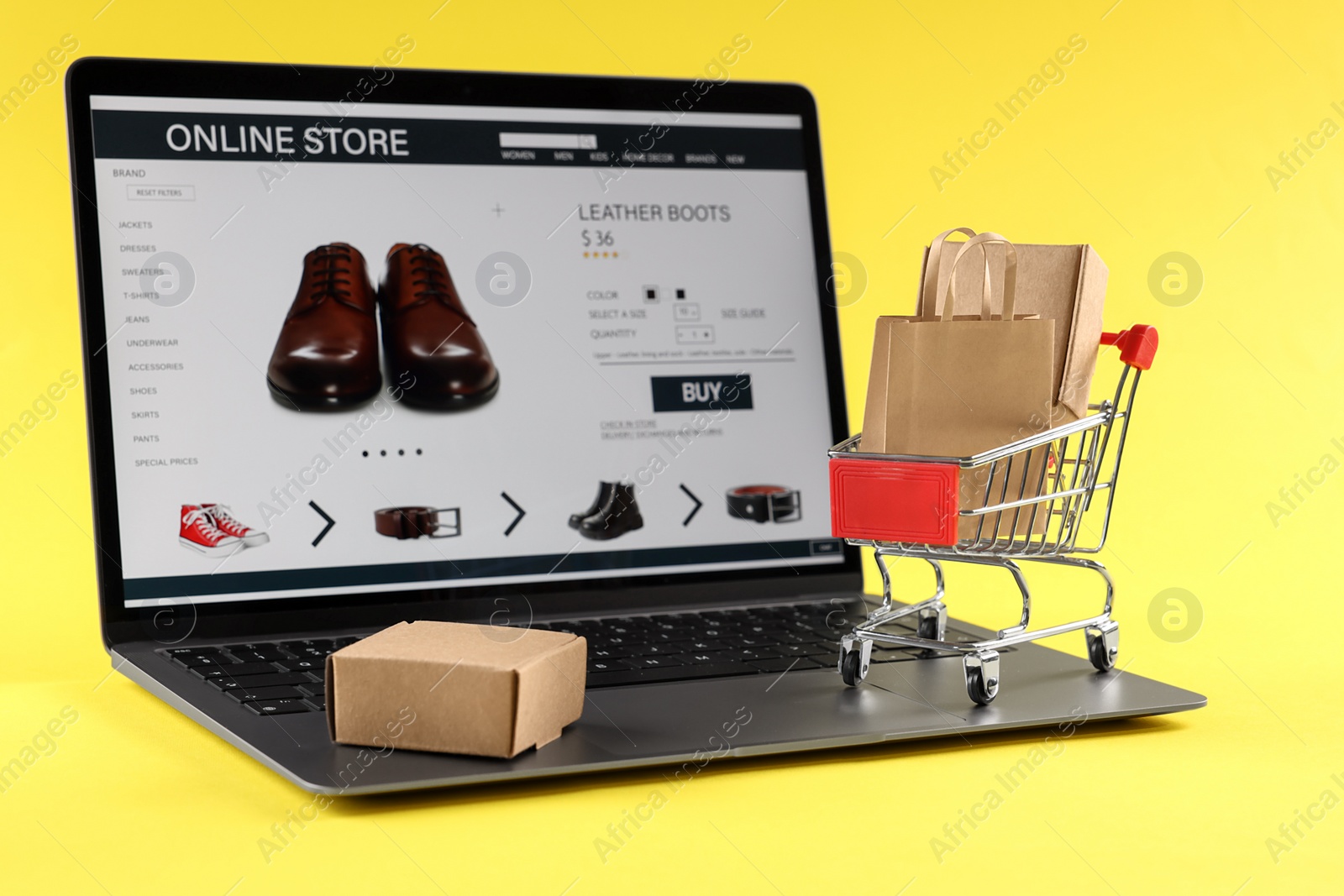 Photo of Online store. Laptop, mini shopping cart and purchases on yellow background