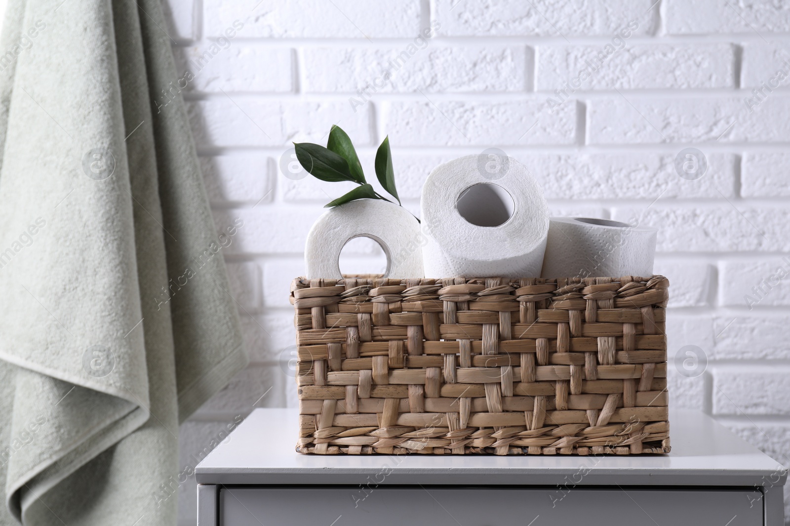 Photo of Toilet paper rolls in wicker basket and floral decor on chest of drawers near white brick wall