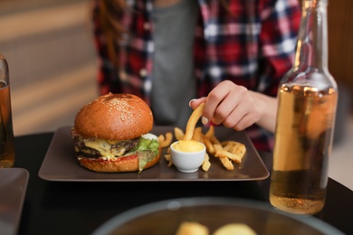 Photo of Young woman eating french fries and tasty burger in cafe, closeup