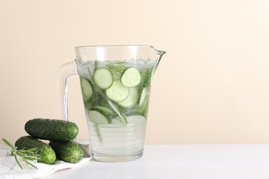 Photo of Refreshing cucumber water with rosemary in jug and vegetables on white table against beige background. Space for text