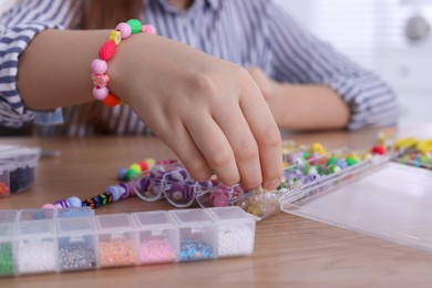 Photo of Little girl making beaded jewelry at table indoors, closeup