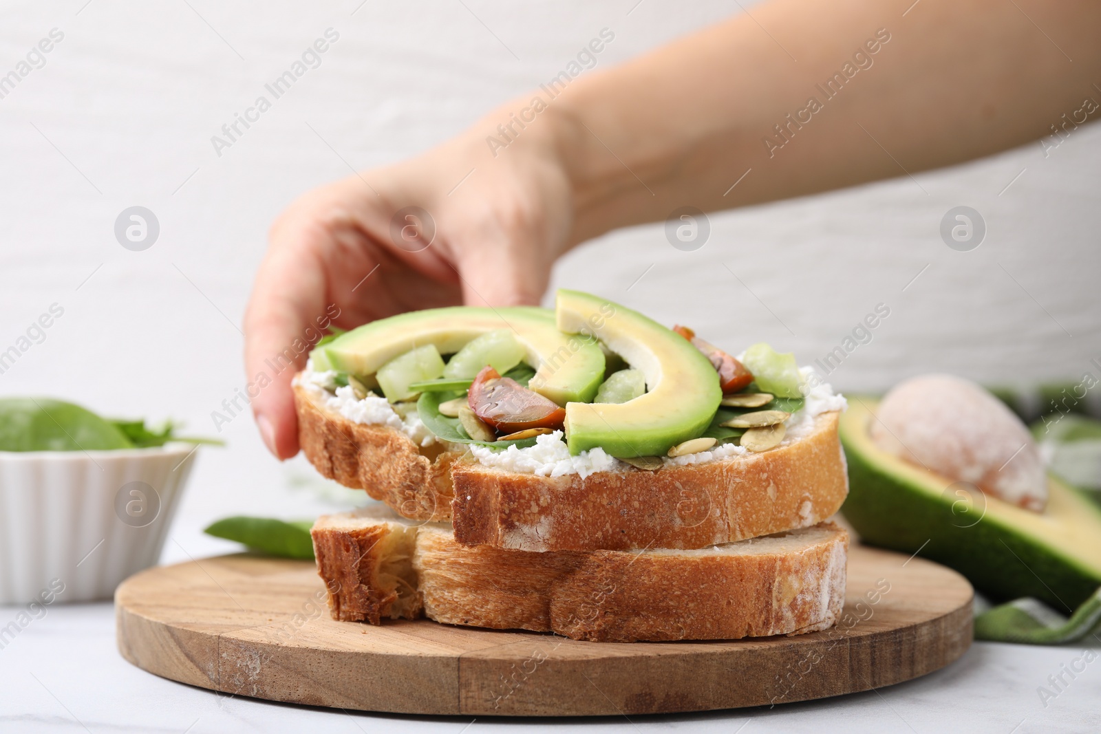 Photo of Woman taking tasty vegan sandwich with avocado, tomato and spinach at white marble table, closeup