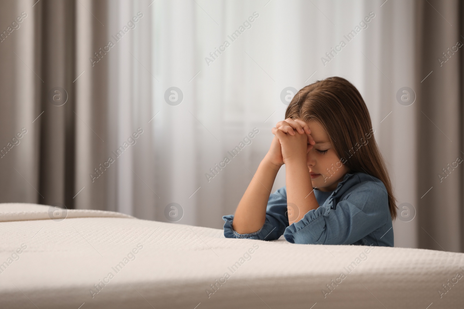 Photo of Cute little girl with hands clasped together saying bedtime prayer in bedroom. Space for text