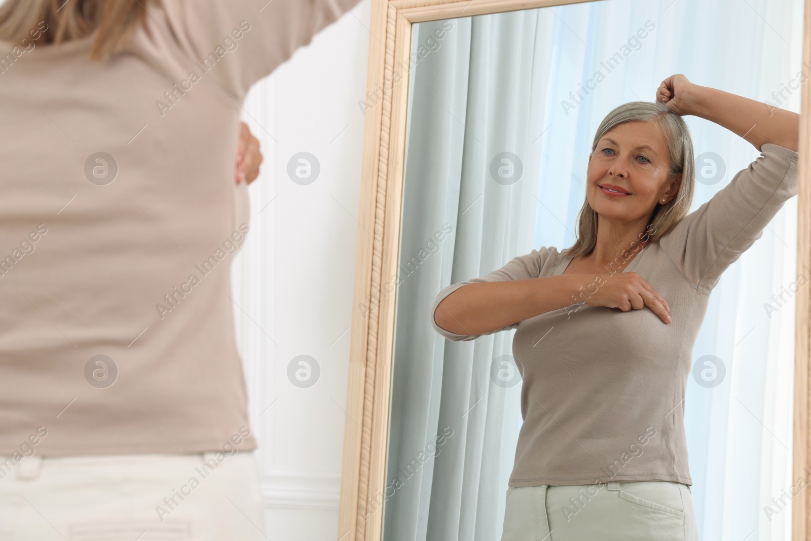 Photo of Beautiful senior woman doing breast self-examination near mirror indoors