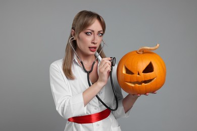 Emotional woman in scary nurse costume with carved pumpkin and stethoscope on light grey background. Halloween celebration