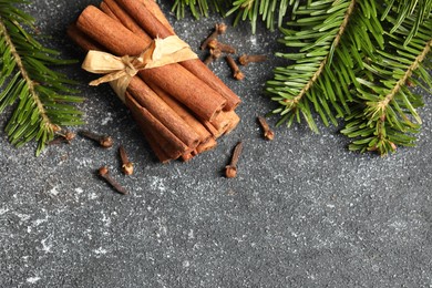 Photo of Different spices. Aromatic cinnamon sticks, clove seeds and fir branches on dark gray textured table, flat lay. Space for text