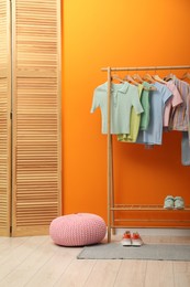 Rack with different stylish women's clothes, shoes and pouf near orange wall indoors