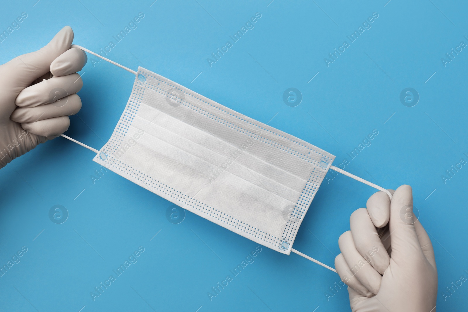 Photo of Doctor in medical gloves holding protective mask on light blue background, closeup. Space for text