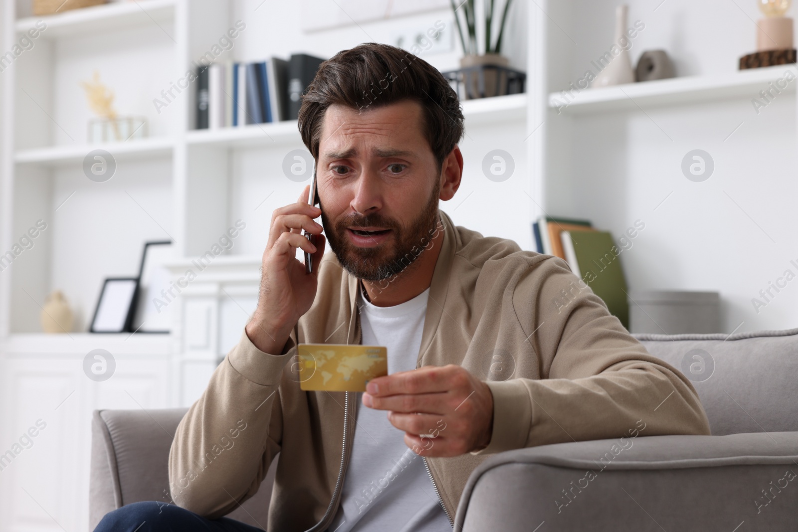 Photo of Upset man with credit card talking on phone in armchair at home. Be careful - fraud