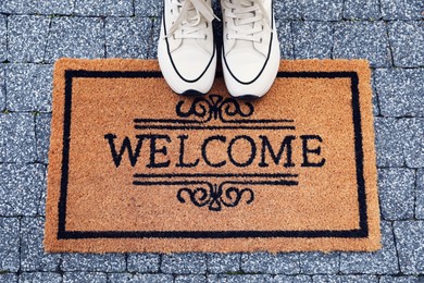 Doormat with word Welcome and stylish sneakers on pavement outdoors, flat lay