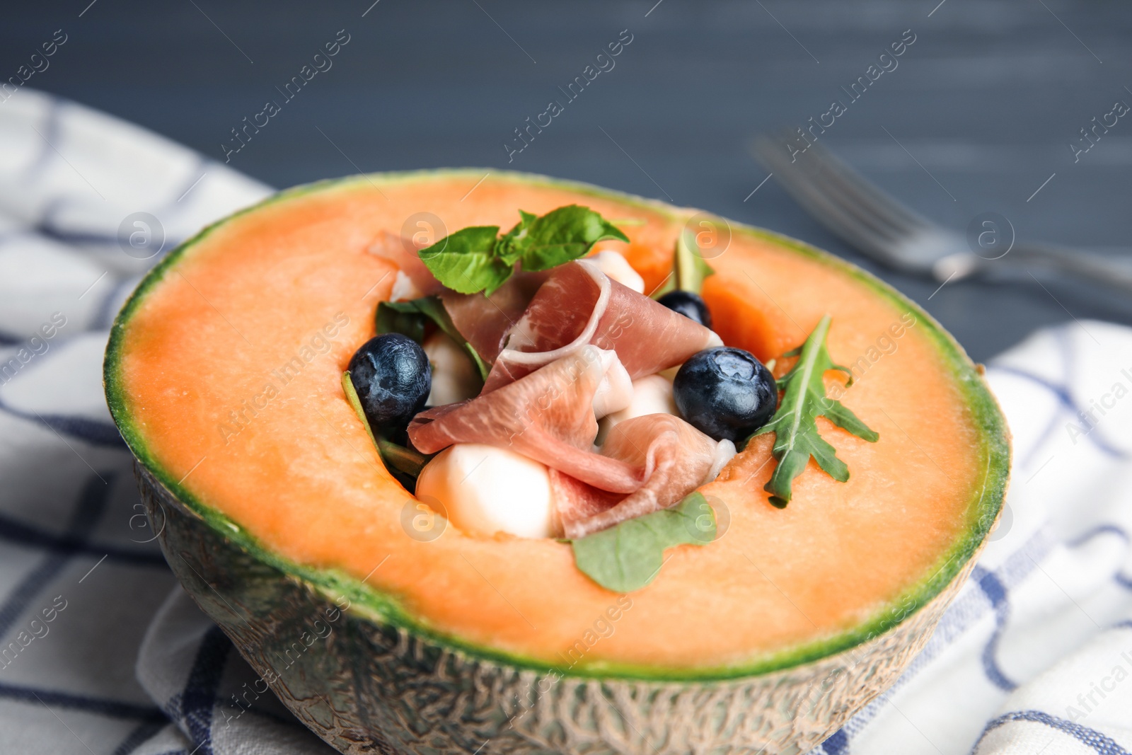 Photo of Half of melon with prosciutto, blueberries and herbs on table, closeup