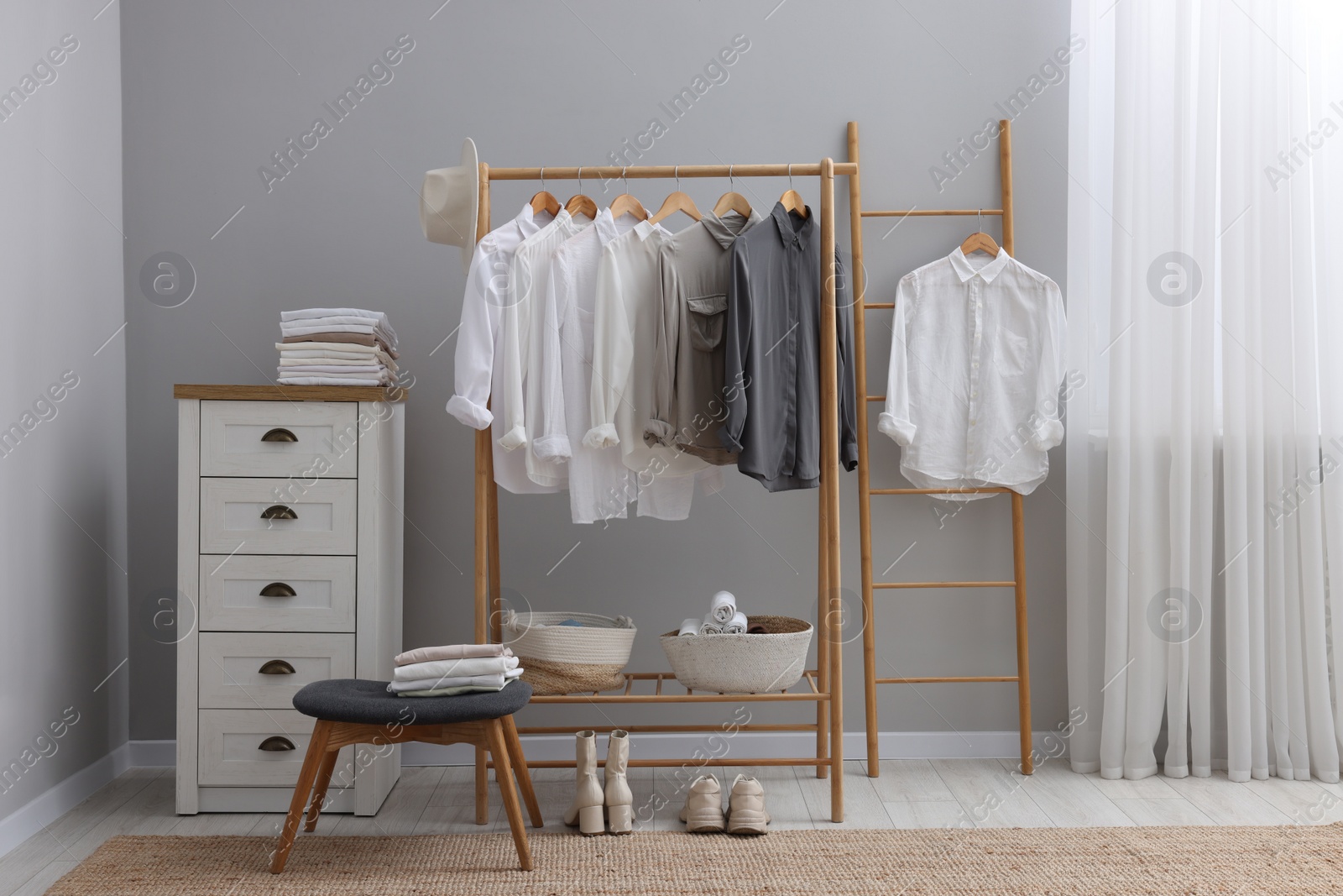 Photo of Wardrobe organization. Rack with different stylish clothes, ottoman and chest of drawers near grey wall in room