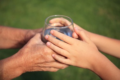 Photo of Child giving glass of water to elderly woman outdoors on sunny day, closeup