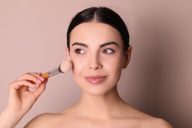 Photo of Beautiful young woman applying face powder with brush on dusty rose background