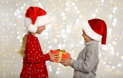 Photo of Happy little children in Santa hats with gift box against blurred festive lights. Christmas celebration